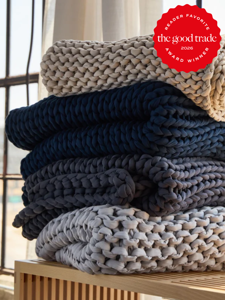 Three chunky knit blankets in dark blue, gray, and beige are stacked on a wooden surface near a window. A red "The Good Trade Reader Favorite 2026 Award Winner" badge is in the top right corner.