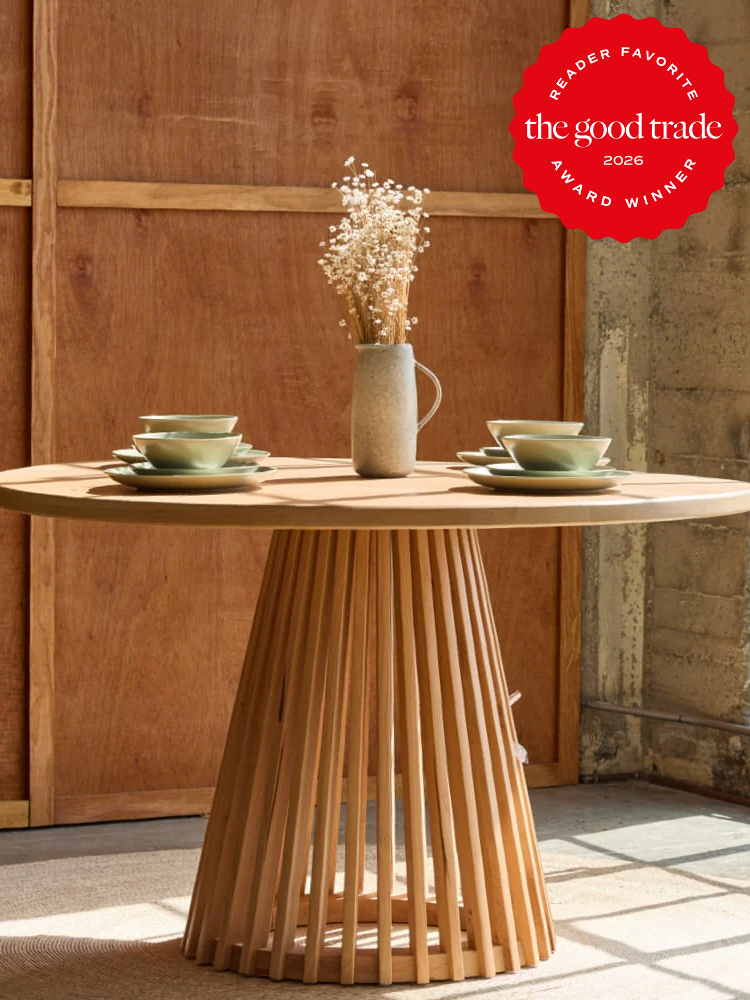 Round wooden table with a slatted base, set with green dishes and a beige vase holding dried flowers; "the good trade 2026 award winner" badge in the upper right corner.