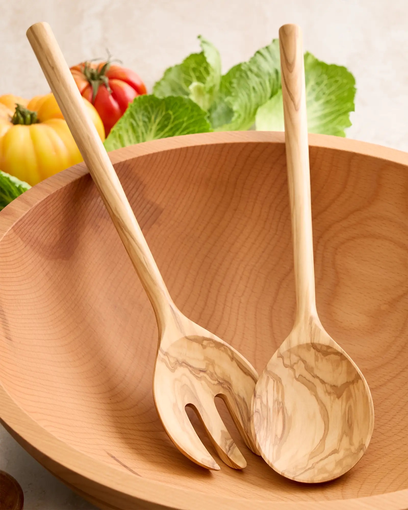 Two wooden salad servers, one spoon and one fork, rest inside a wooden bowl with tomatoes and leafy greens in the background.