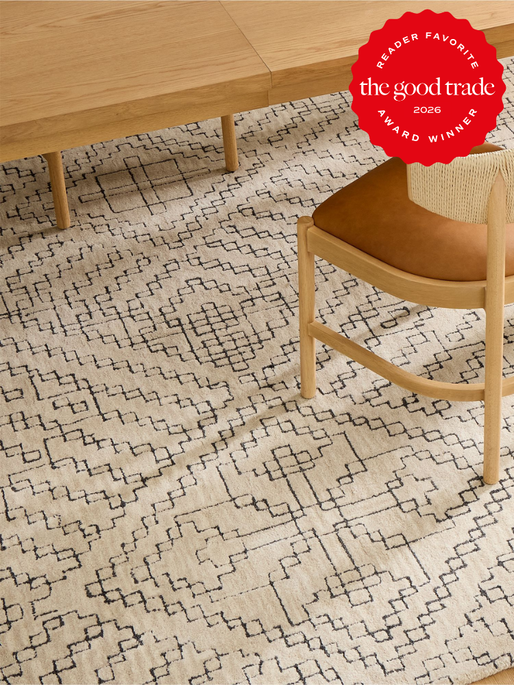 A light wooden table and chair with tan leather seat rest on a beige rug with a geometric black pattern; a red "The Good Trade Award Winner 2026" badge is in the top right corner.