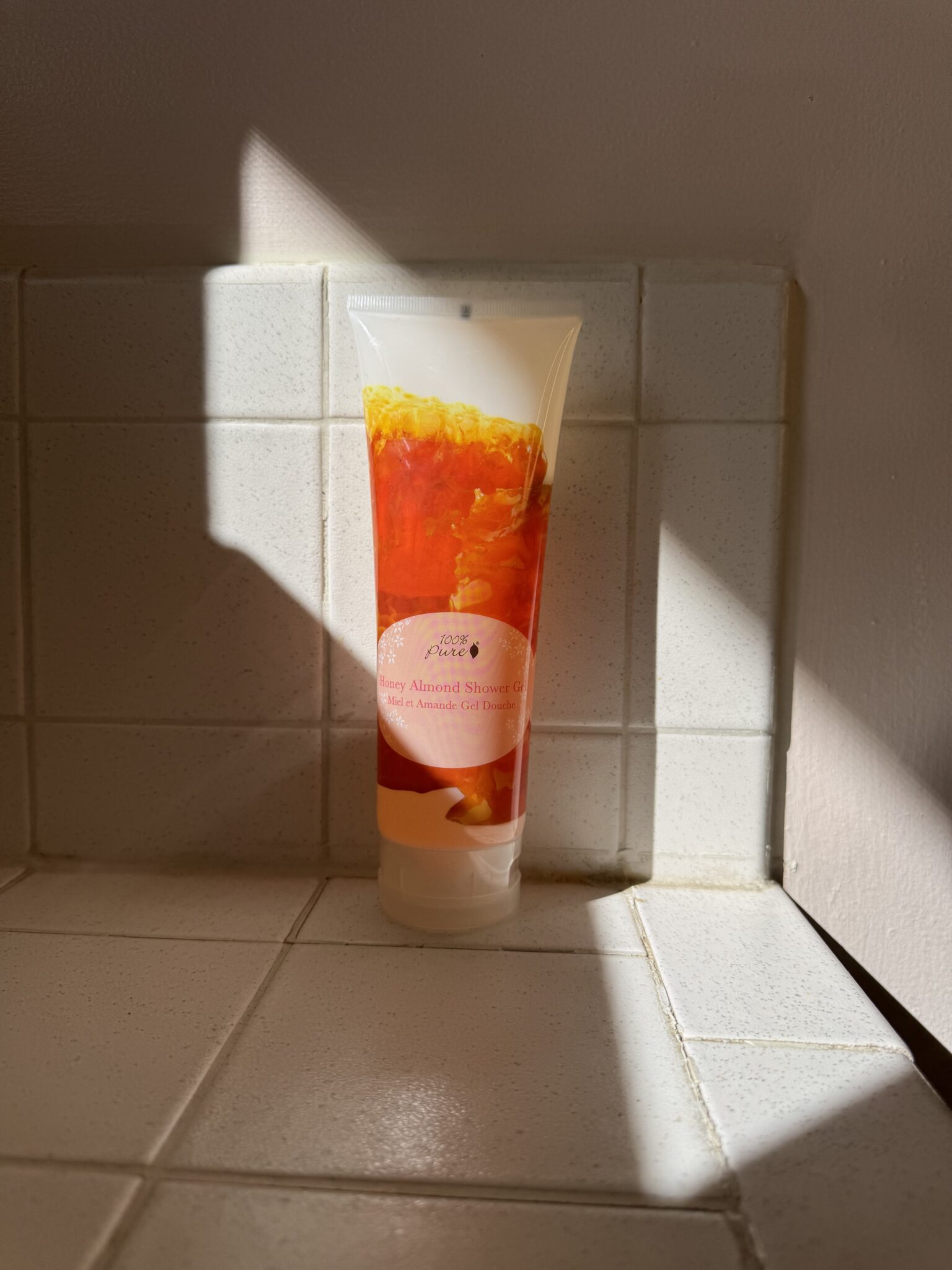 A tube of shower gel with an orange floral design stands upright on a tiled bathroom surface, illuminated by a shaft of sunlight.