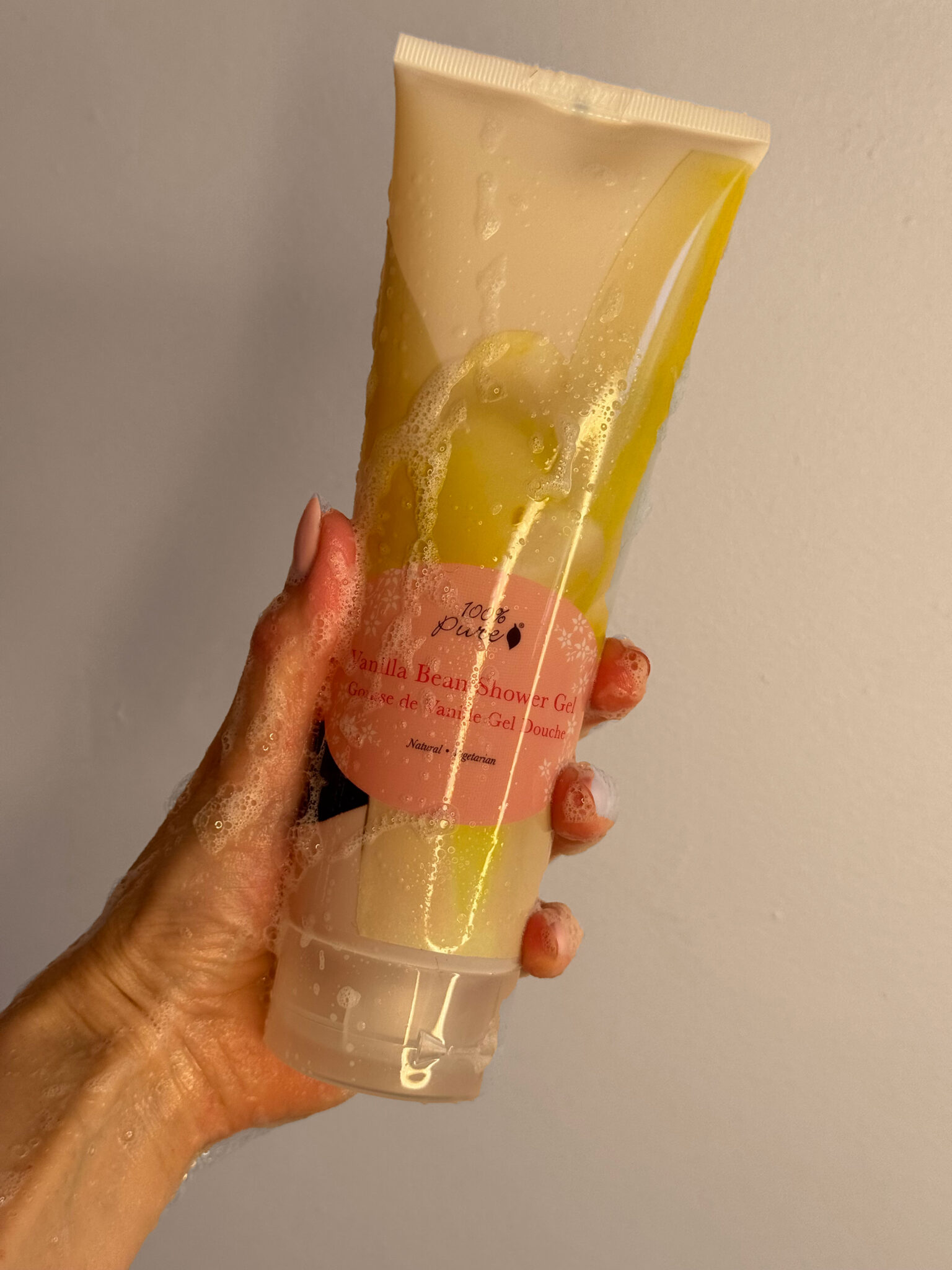 A hand holding a bottle of shower gel with a pink label, covered in water droplets against a plain background.