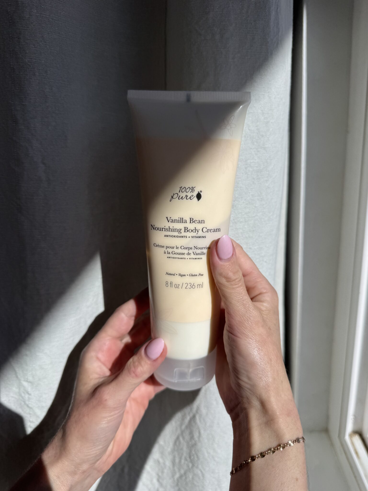 A person holds a tube of 100% Pure Vanilla Bean Nourishing Body Cream near a window with sunlight shining on their hands.
