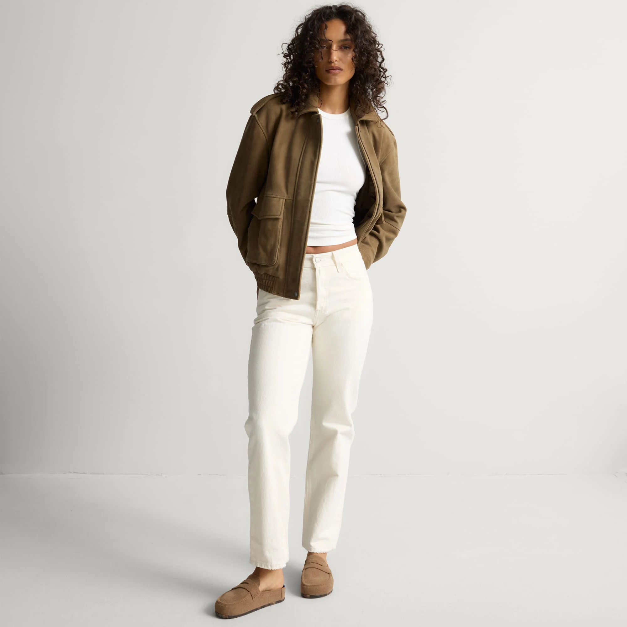 Person standing against a plain background, wearing a brown jacket, white t-shirt, white pants, and brown shoes.