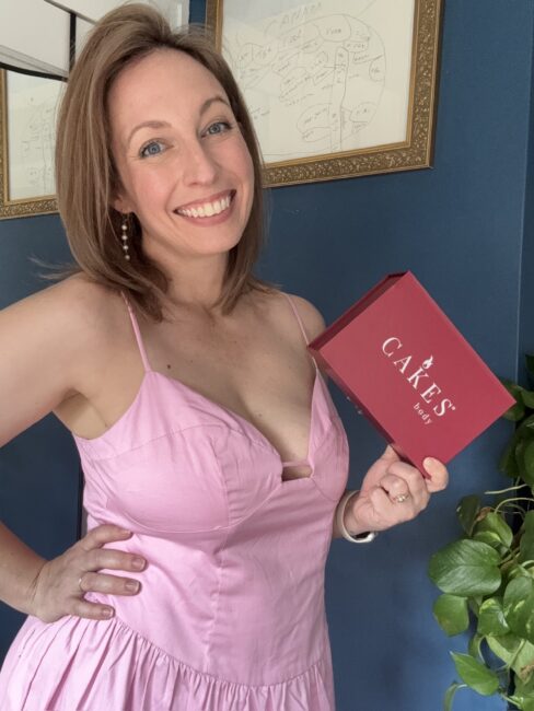Woman in a pink dress smiles and holds a red box labeled "Cakes" in front of a dark blue wall with a framed map and a green plant.