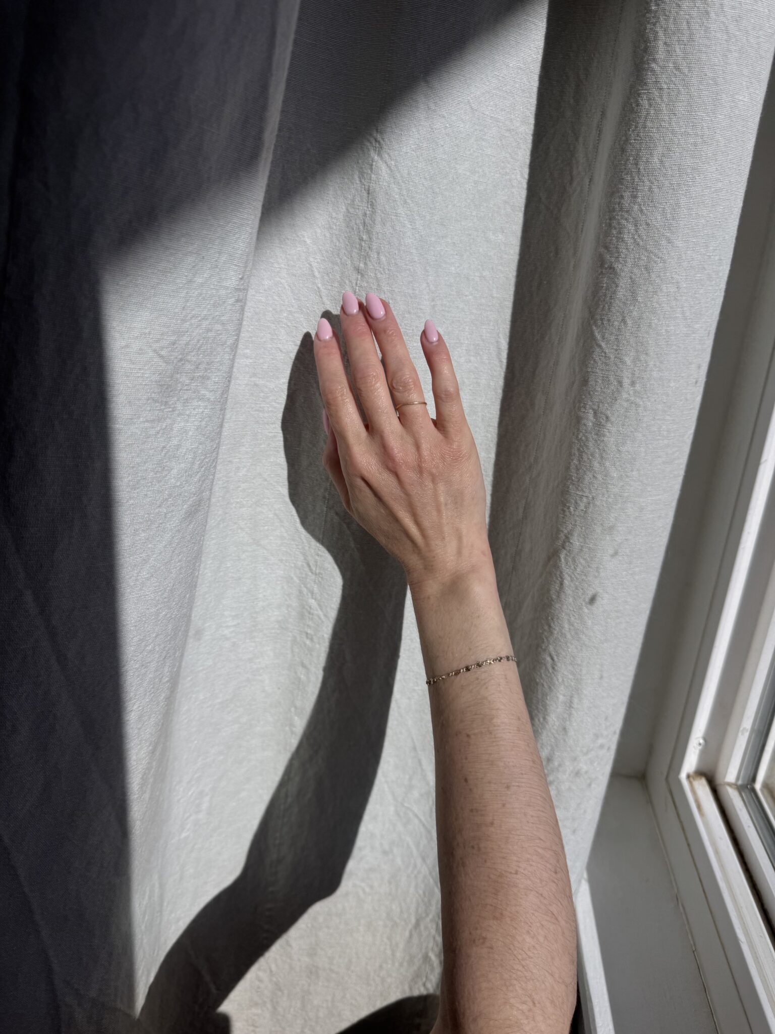 A hand with light pink nails touches a sunlit white curtain, casting a shadow on the fabric near a window.