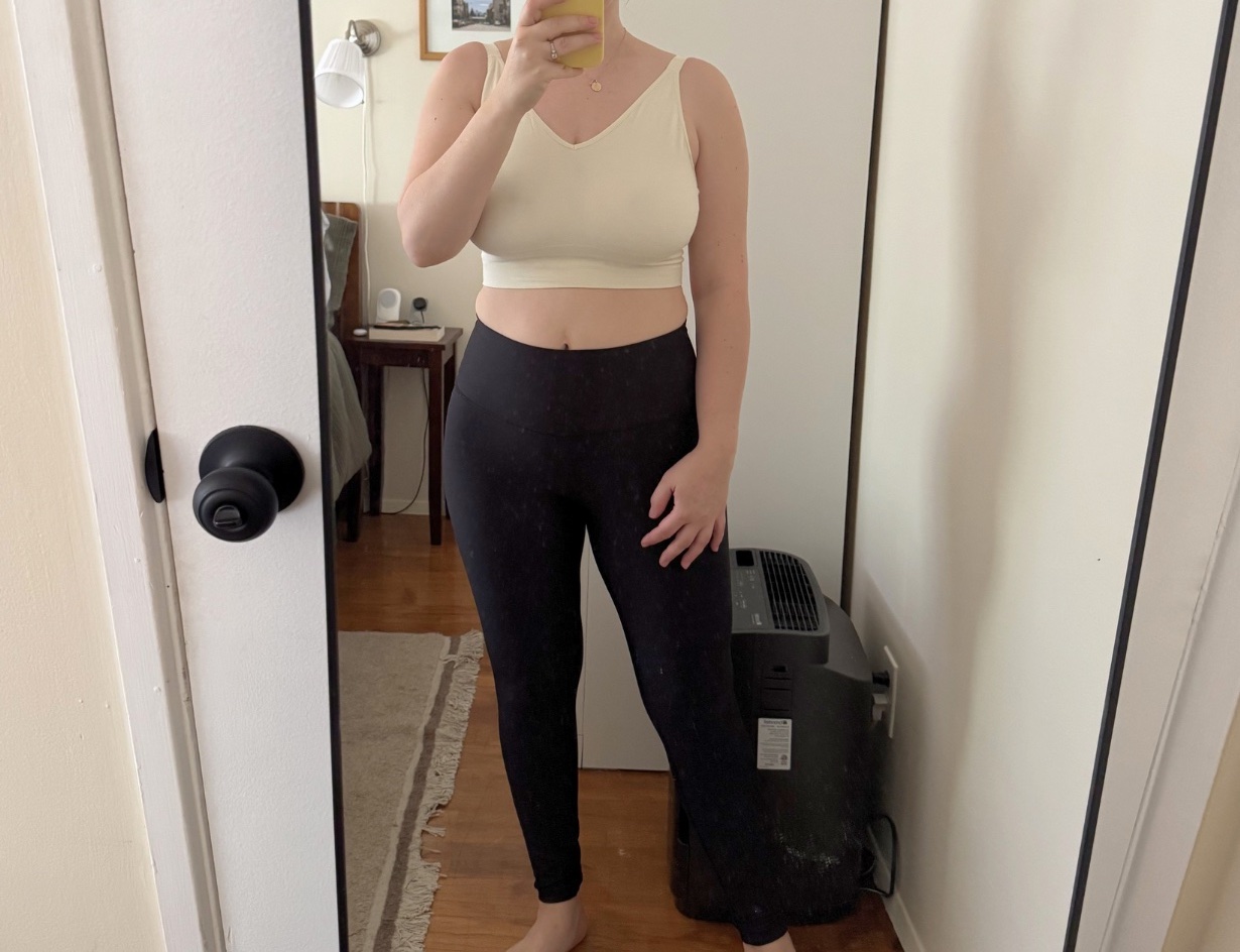 A person stands barefoot in front of a mirror wearing a white crop top and black leggings, with one hand on their hip. A room with a bed and small heater is visible in the background.