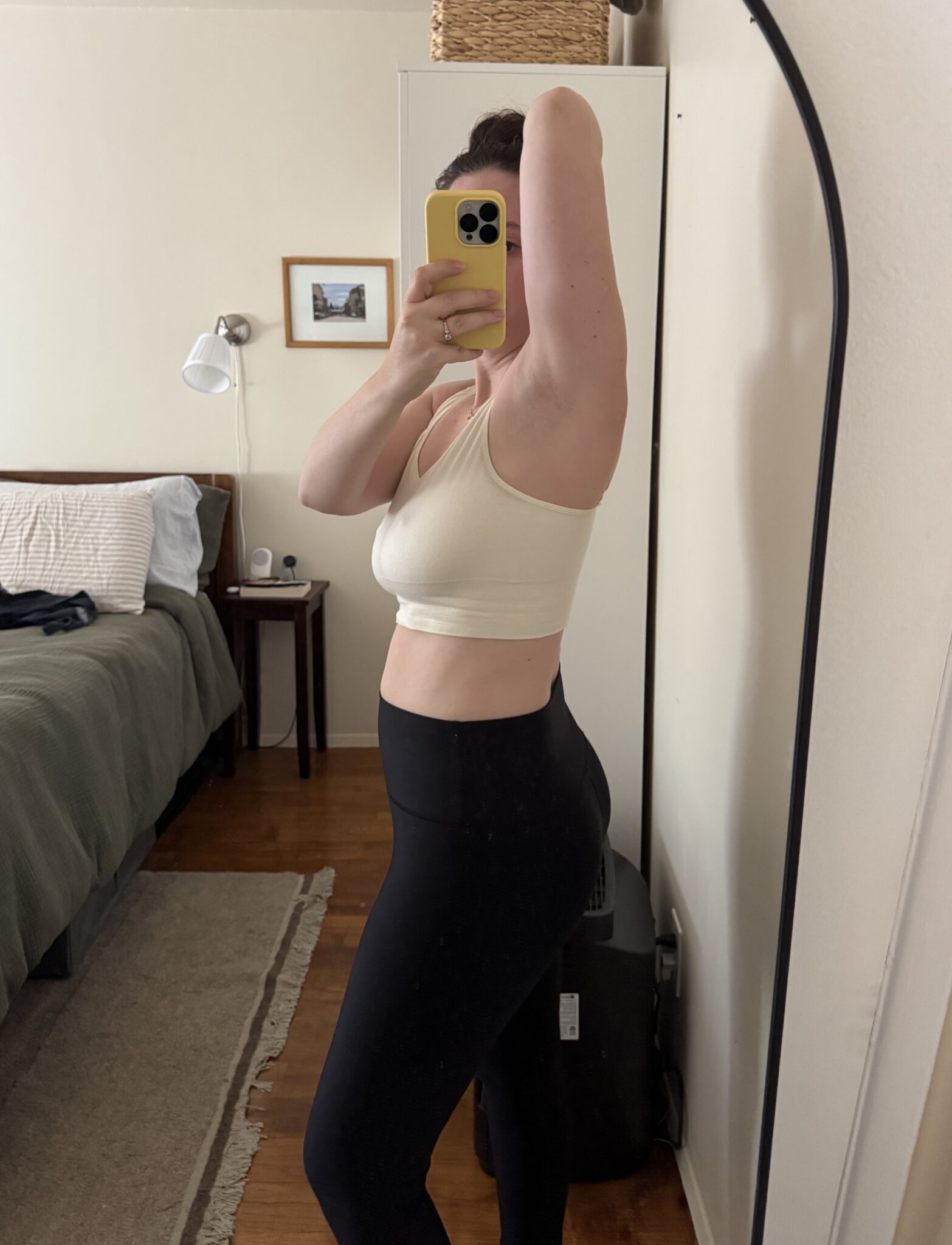 Person wearing a white crop top and black leggings takes a side-view mirror selfie in a bedroom, with arm raised, partially covering their face with a yellow phone.