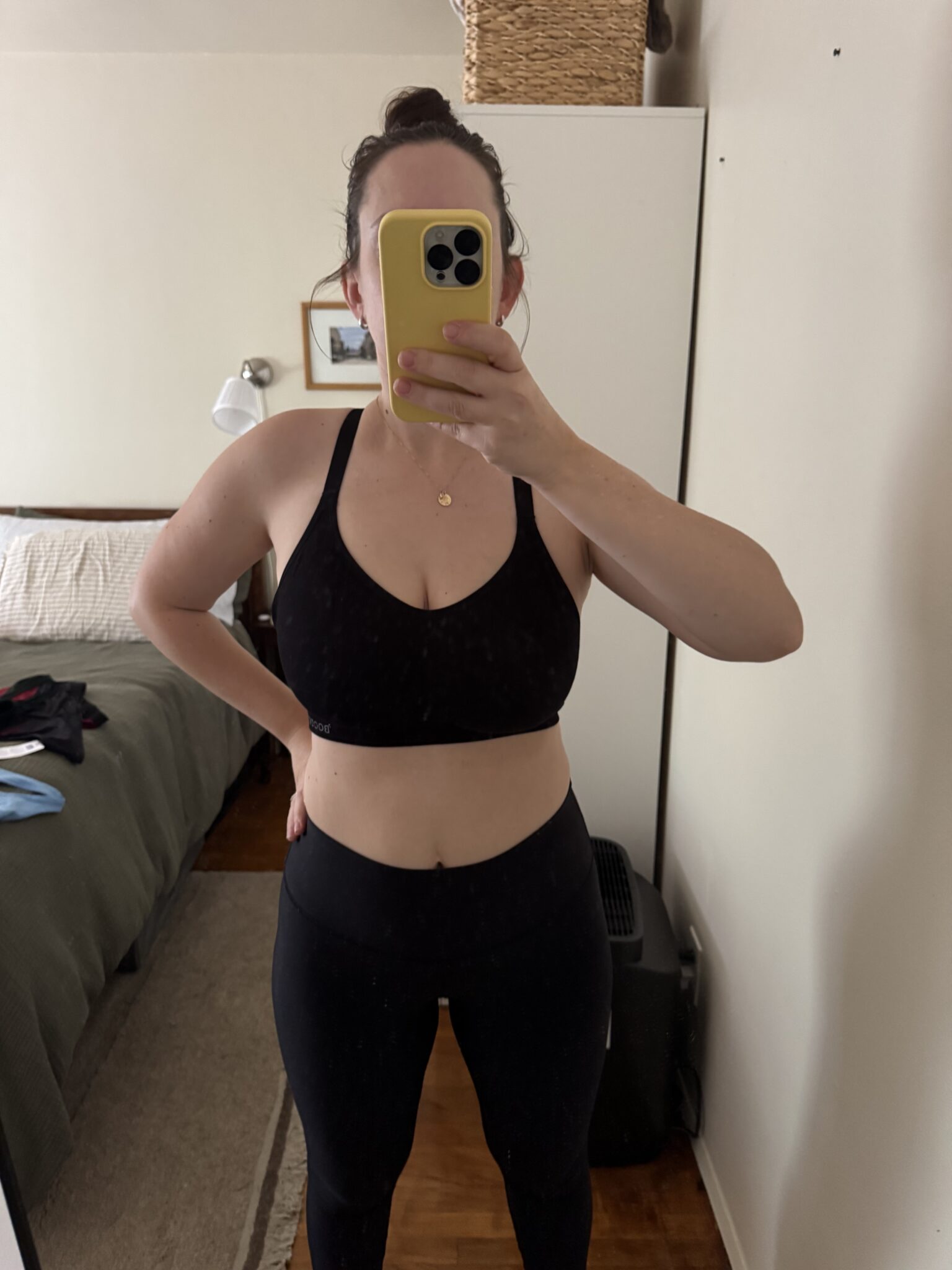 Person wearing a black sports bra and leggings takes a mirror selfie in a bedroom, partially obscuring their face with a yellow phone.