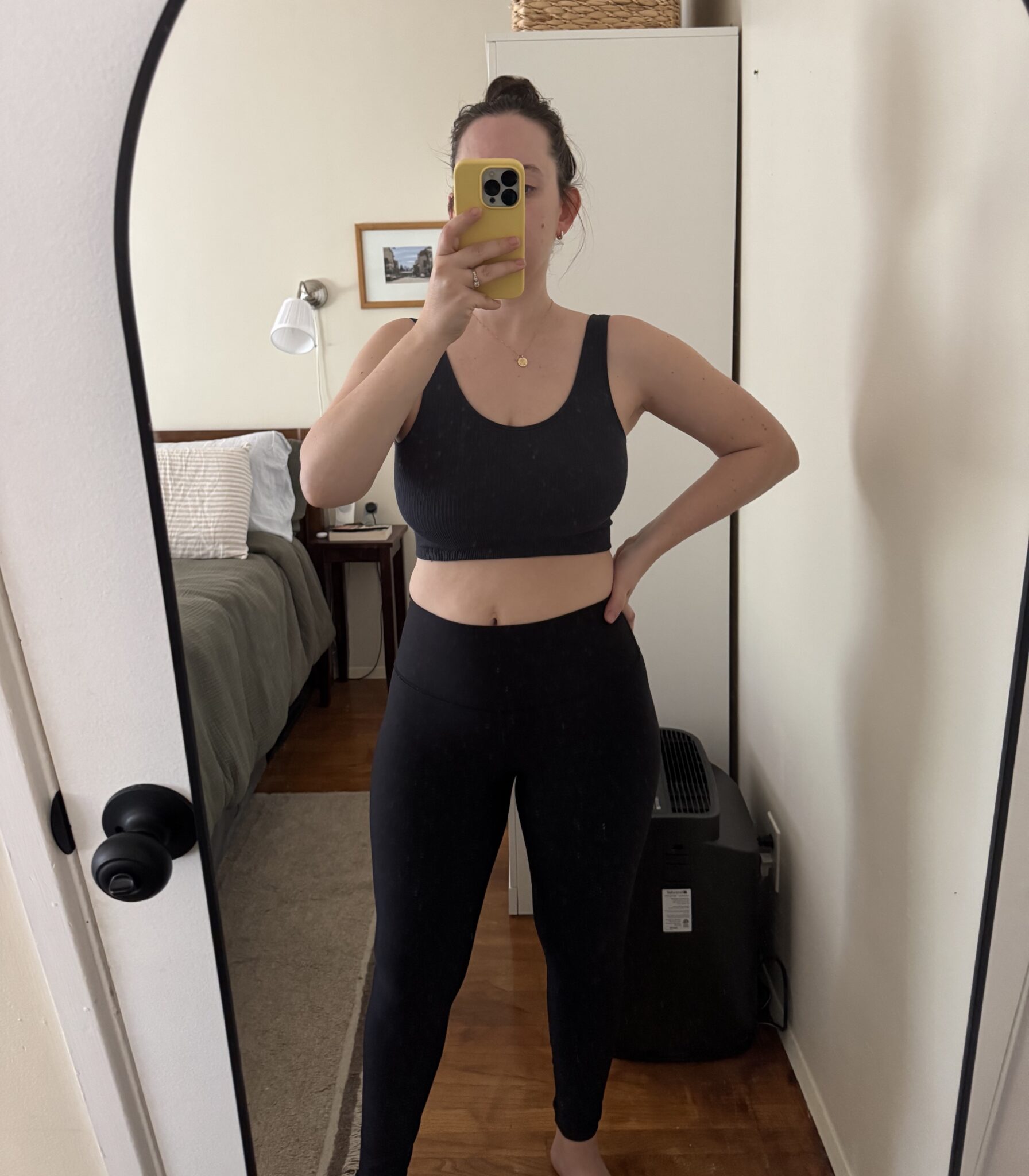 A person in black workout clothes stands in front of a mirror, taking a selfie in a bedroom with a bed, lamp, and wardrobe visible in the background.