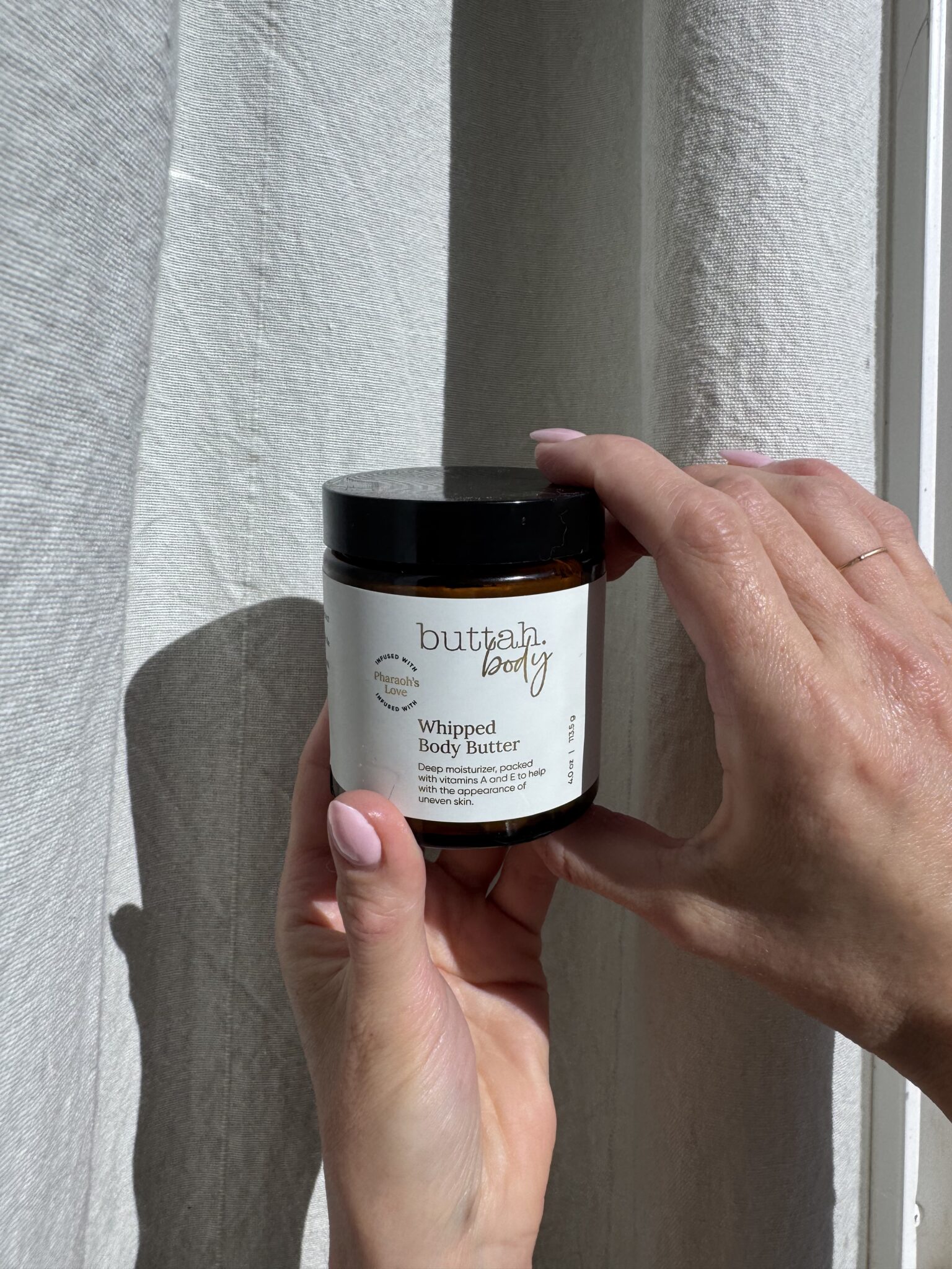 A hand holds a jar of Buttah Body Whipped Body Butter in front of a light-colored curtain, with sunlight casting shadows on the jar and hands.