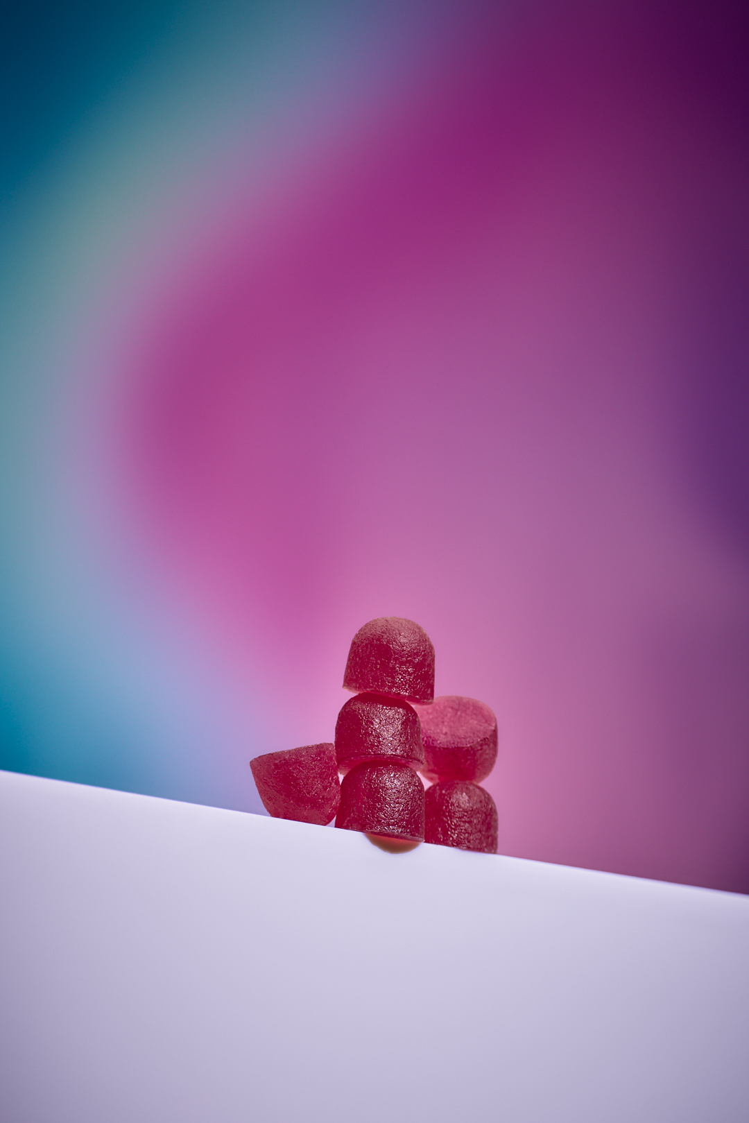 Six red gummy candies are stacked on a white surface with a blurred pink and blue gradient background.
