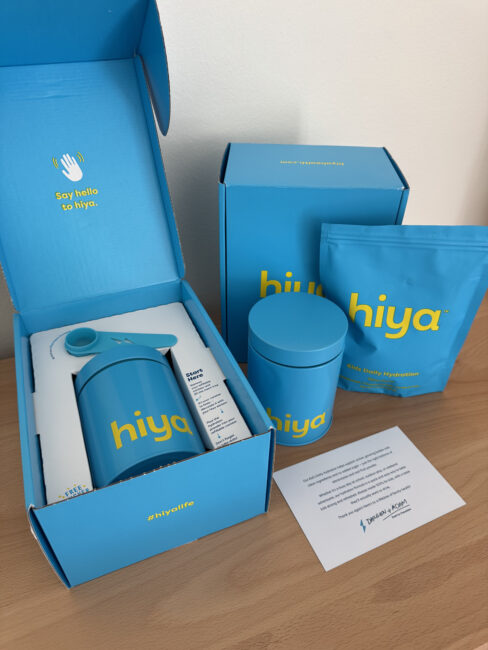A blue Hiya branded water bottle, container, packet, and info card are displayed on a wooden surface with open blue packaging.