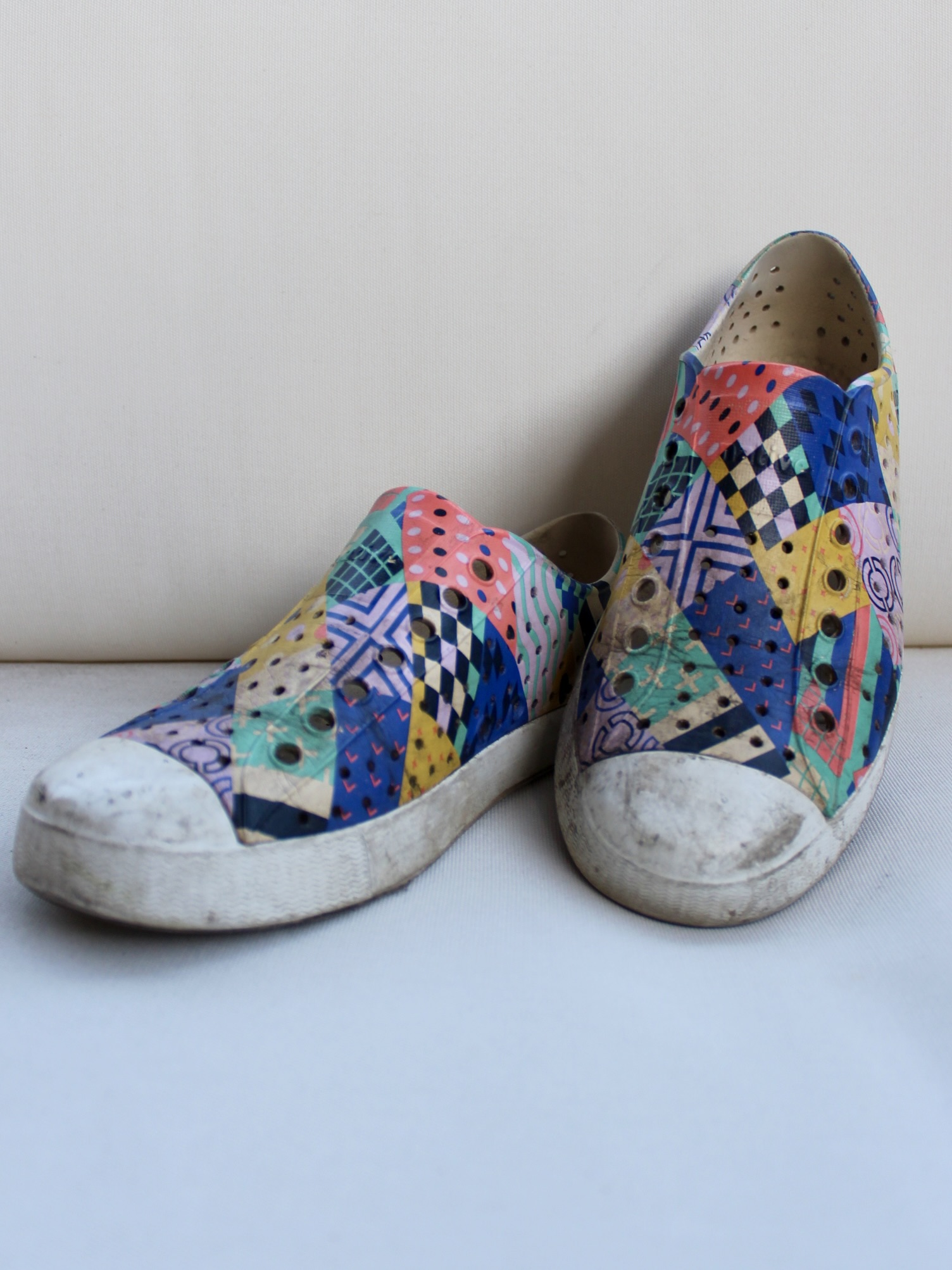 A pair of sustainable sneakers in slip-on canvas style, featuring a colorful geometric patchwork pattern and visible signs of wear on the soles and toes.