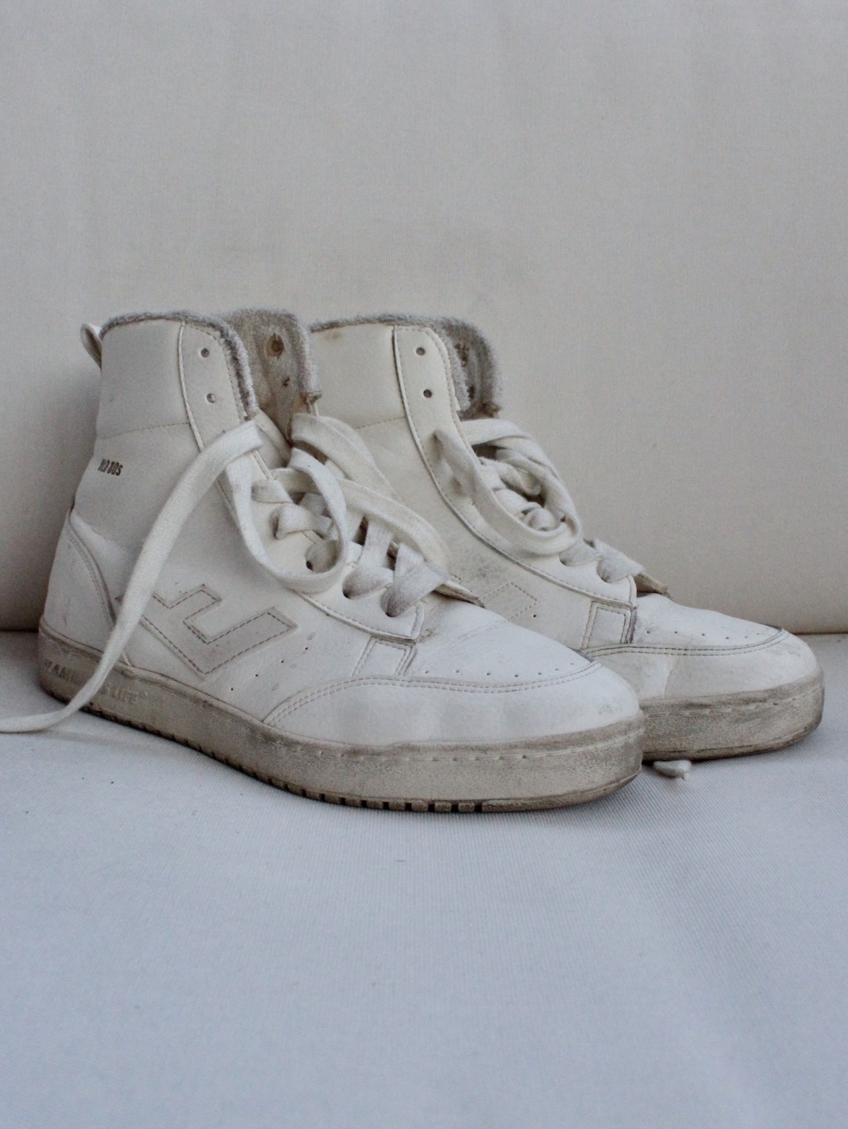 A pair of worn white high-top sustainable sneakers with visible scuff marks and one untied lace, placed on a light surface against a neutral background.