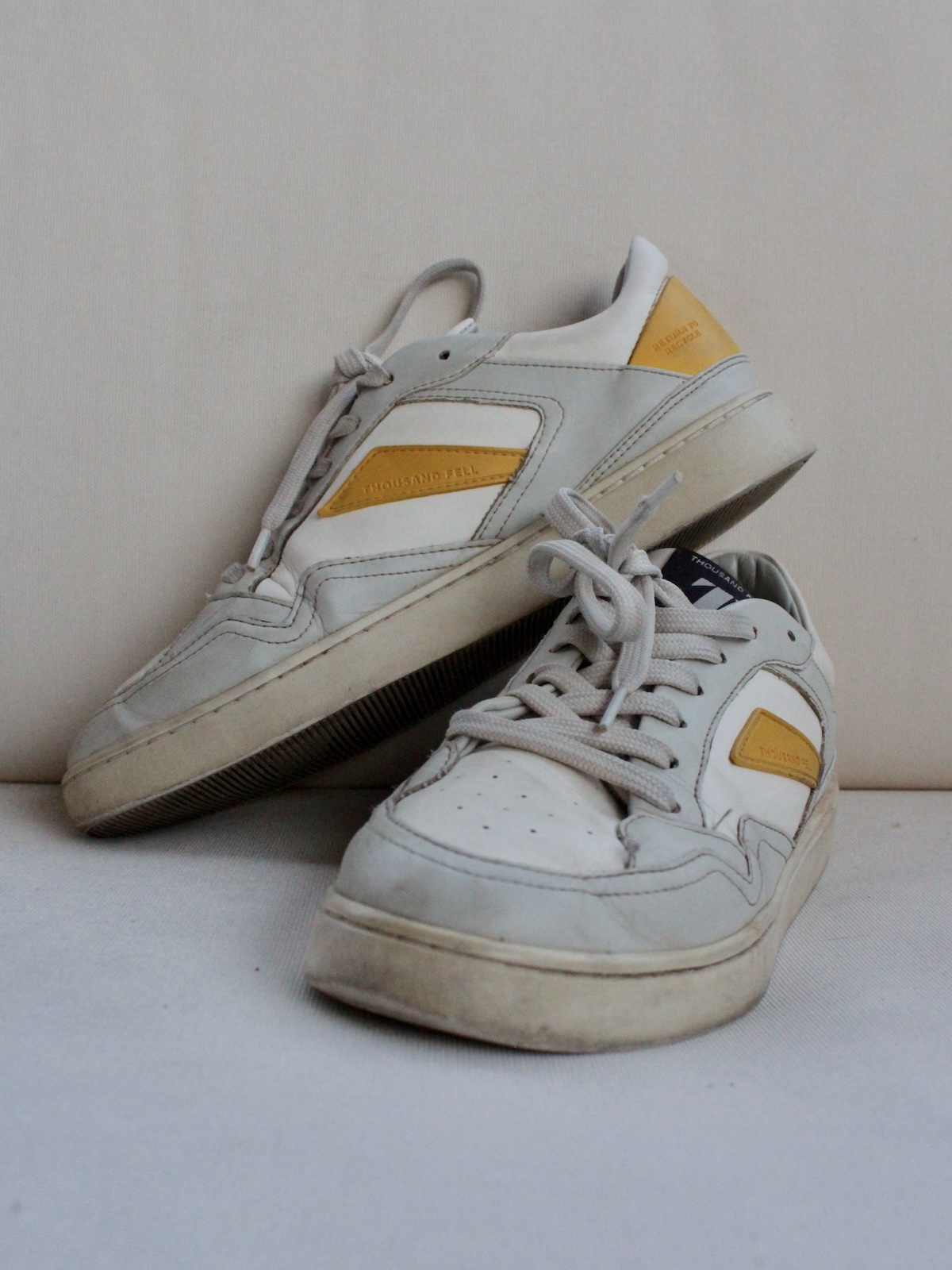 A pair of worn sustainable sneakers with white and yellow accents displayed against a plain light-colored background.