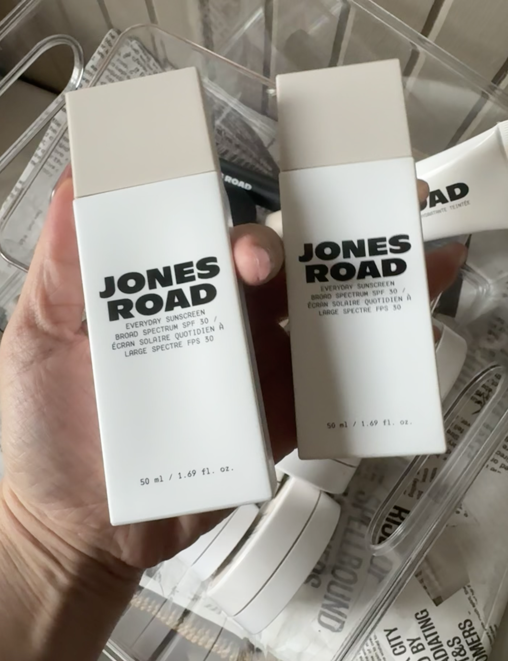 A hand holds two bottles of Jones Road Everyday Sunscreen SPF 30, 50 ml, with Ilia and other skincare products along with a newspaper visible in the background.