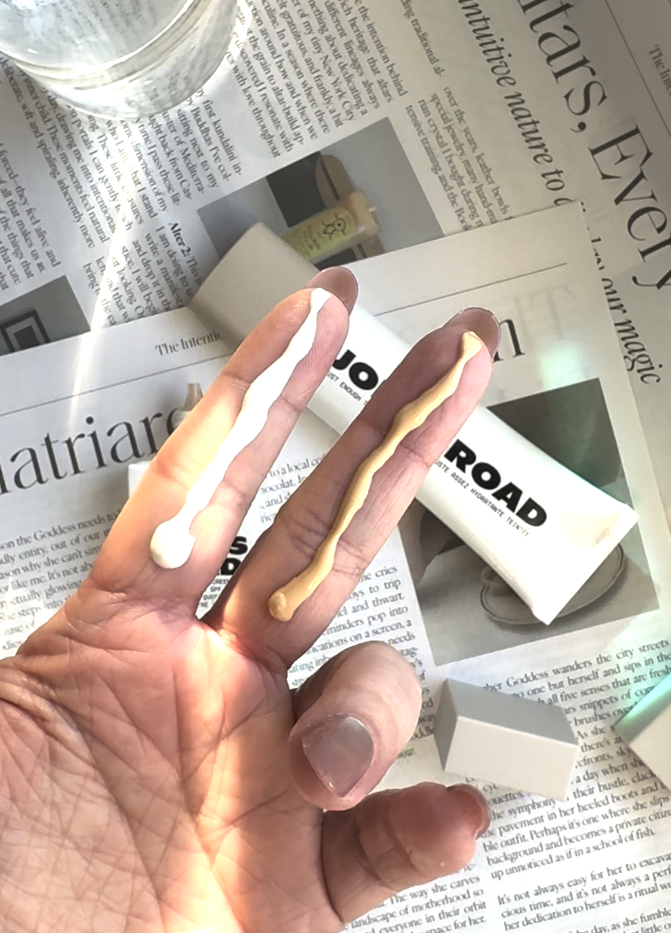 A hand holds up two fingers, each with a line of Ilia cream—one white, one beige—in front of an open skincare tube resting on newspaper pages.