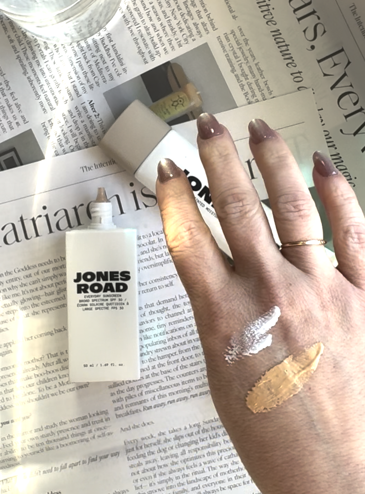 A hand with two swatches of makeup, including a shade from Ilia, rests on a newspaper next to an open "Jones Road" bottle. A glass of water and a makeup applicator are also visible.