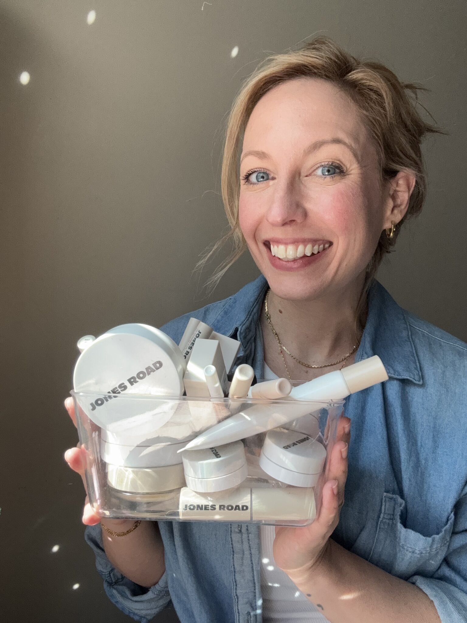A woman smiles at the camera while holding a clear container filled with Jones Road and Ilia beauty products.