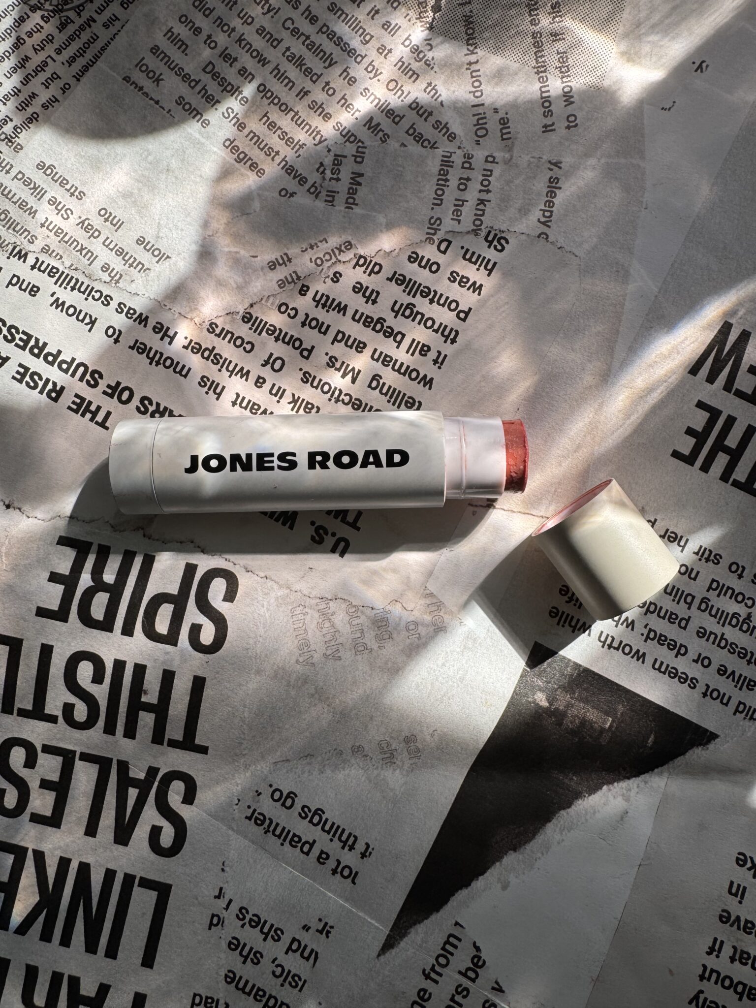 A Jones Road lip balm with its cap off rests on a sheet of printed newspaper under dappled sunlight, reminiscent of an Ilia beauty moment.