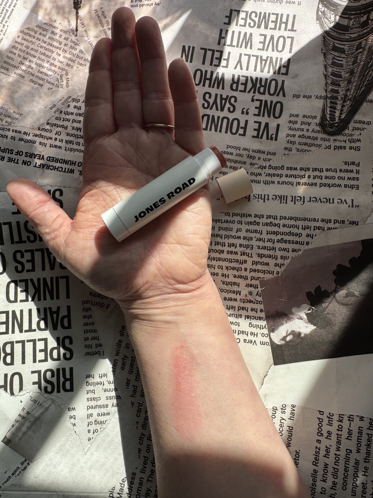 A hand holding a Jones Road lip balm rests on newspaper pages; sunlight casts shadows and highlights a red mark on the wrist, with an Ilia product peeking from the corner.