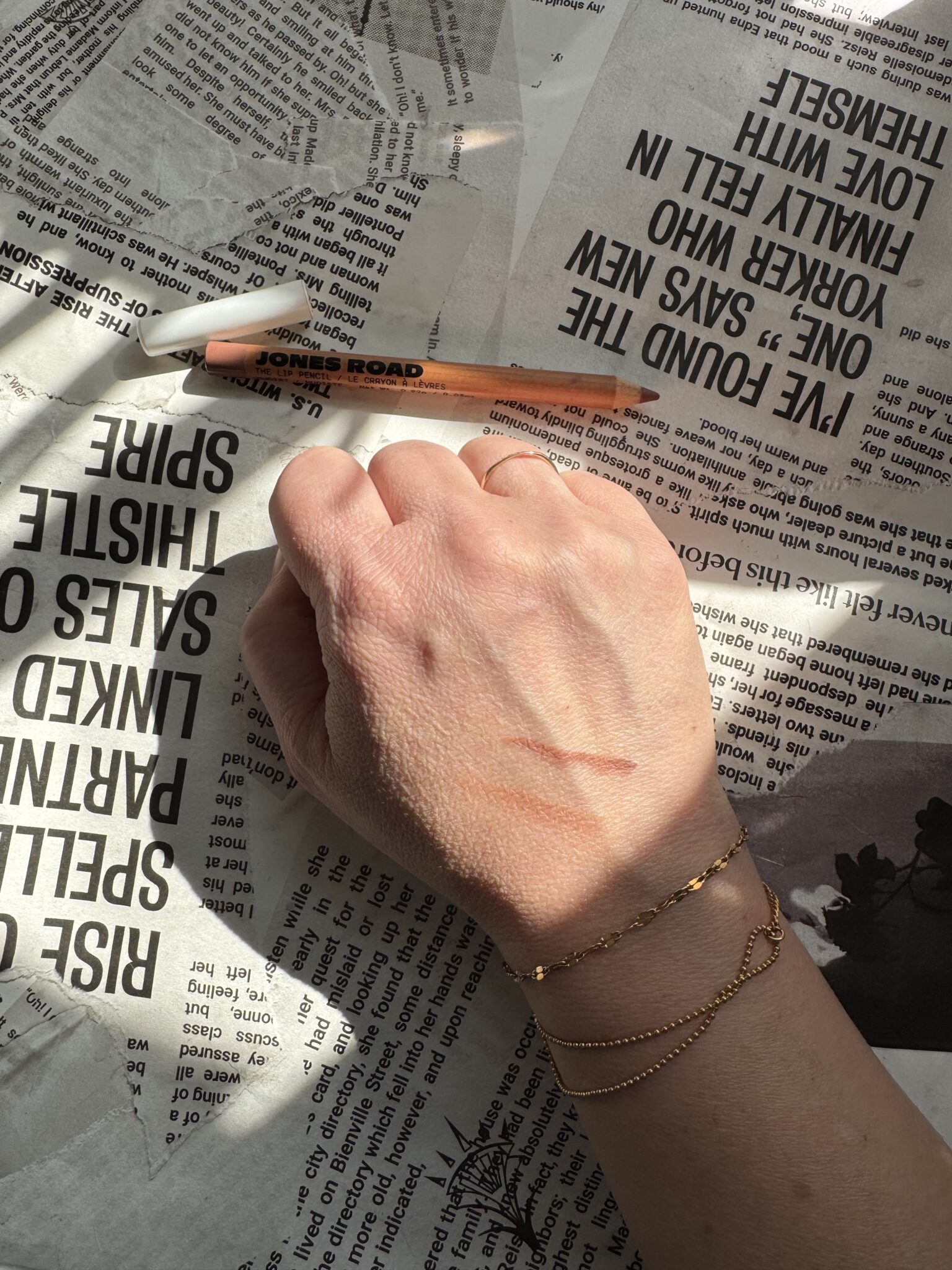 A hand with gold bracelets rests on newspaper, showing two brown makeup swatches on the skin. Nearby lies a brown Jones Road makeup pencil and an Ilia product, adding to the chic beauty lineup.