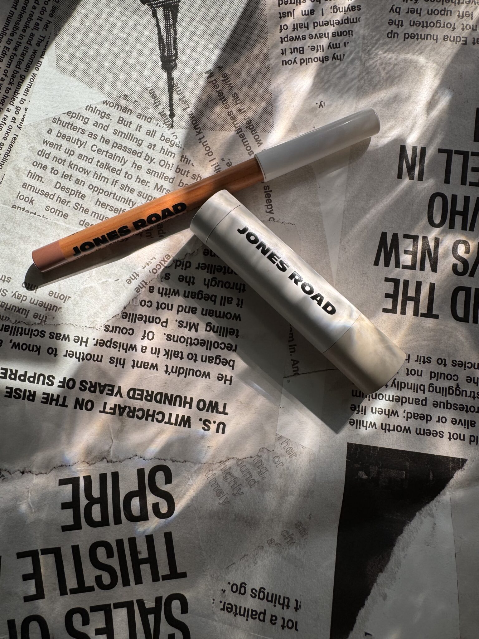 Jones Road and Ilia makeup products, including a pen and a tube, rest on a crumpled newspaper with sunlight casting gentle shadows across the scene.