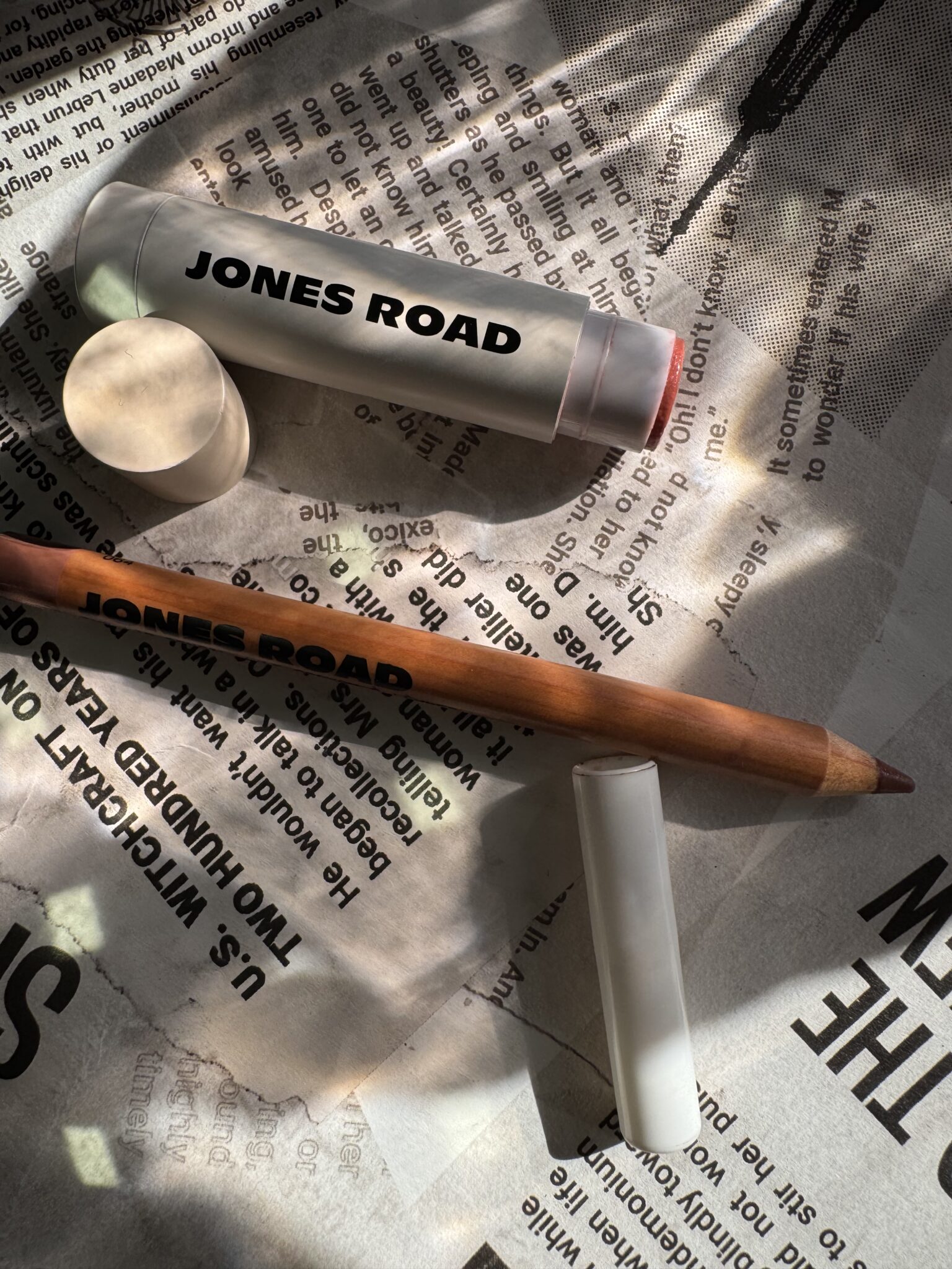 Jones Road lip balm and Ilia lip pencil with their caps off, placed on a newspaper under dappled sunlight.