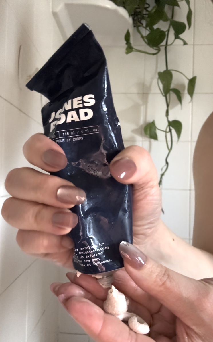 A person squeezes lotion from a dark blue tube labeled "JONES ROAD" onto their hand in a tiled bathroom with a green hanging plant and an Ilia product visible in the background.