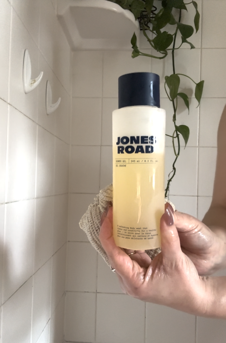 A person holds a bottle of Jones Road foaming face wash in a white-tiled shower, with a plant and towel visible in the background, creating a serene scene reminiscent of Ilia’s clean beauty aesthetic.