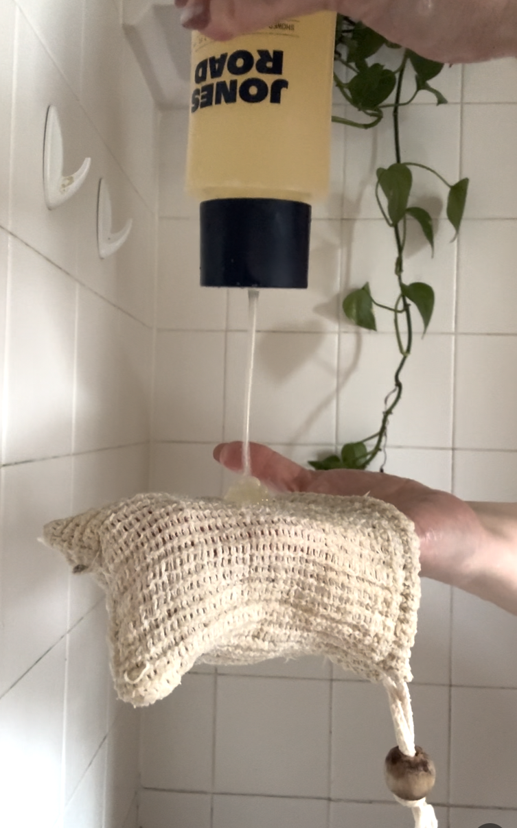 A person dispenses liquid soap from a yellow bottle onto a textured shower loofah in a tiled bathroom with a green vine and an Ilia product on the wall.