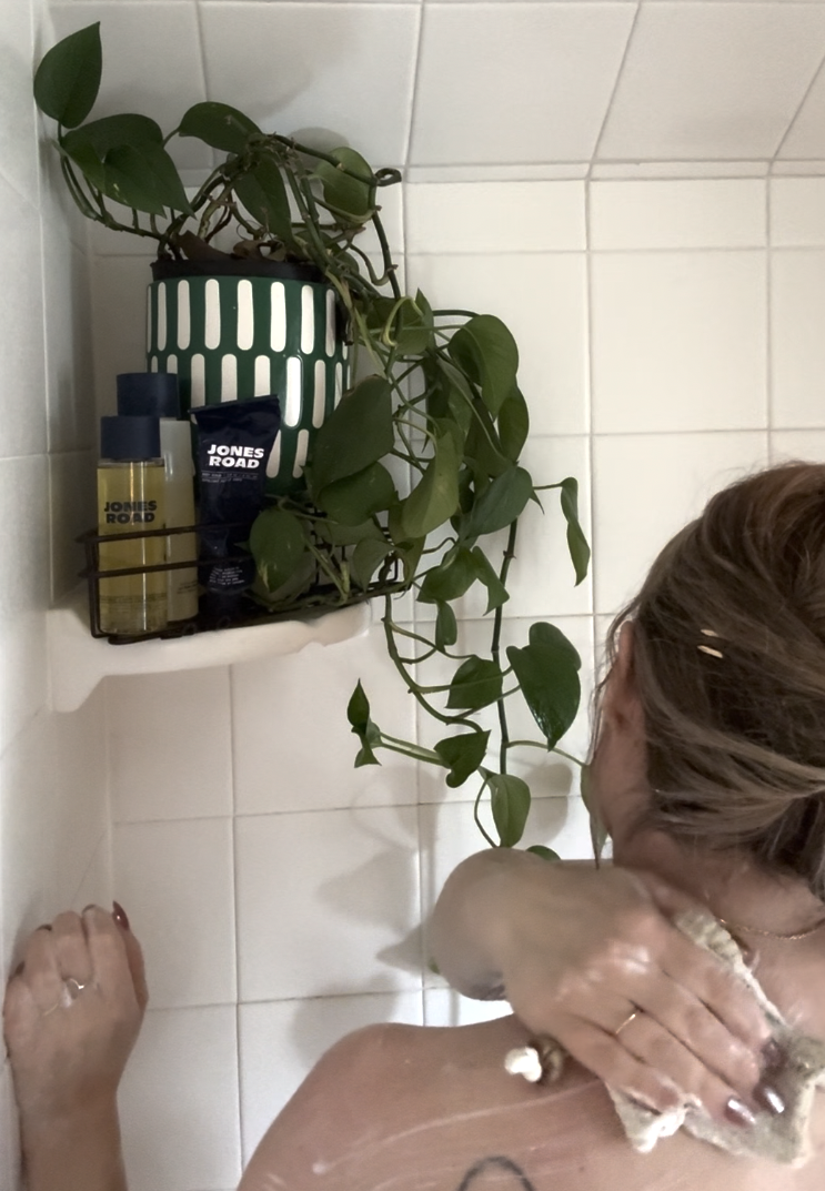 A person washes their upper back in a tiled shower with a plant and Ilia grooming products arranged neatly on a shelf in the corner.