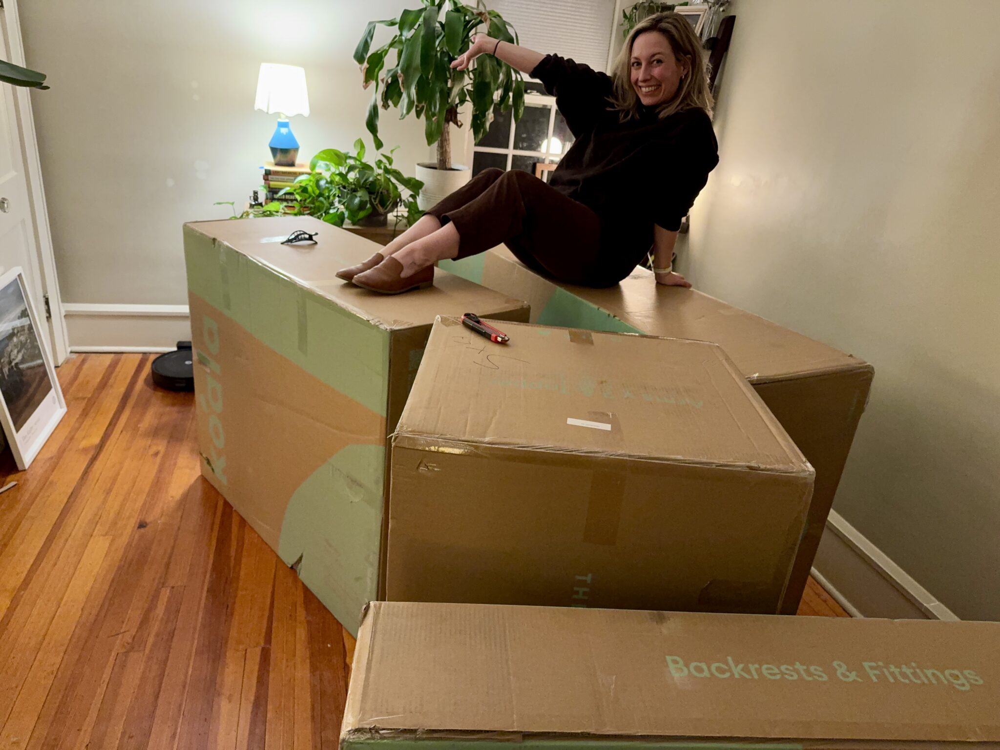 A woman sits and smiles on large cardboard boxes labeled "Backrests & Fittings" in a living room with wooden floors, houseplants, and a lamp in the background.
