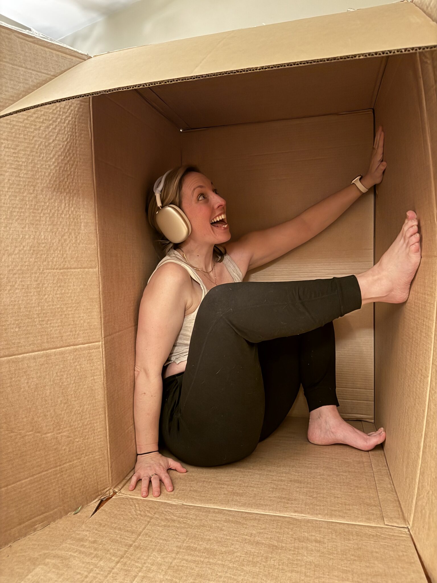 A person wearing headphones sits smiling inside a large cardboard box, with one leg propped against the side and their back against the wall.