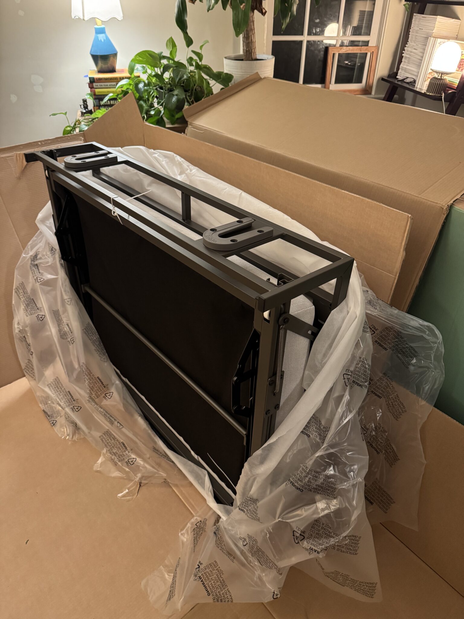 A partially unpacked metal desk frame sits on cardboard and plastic wrap in a room with plants and boxes nearby.