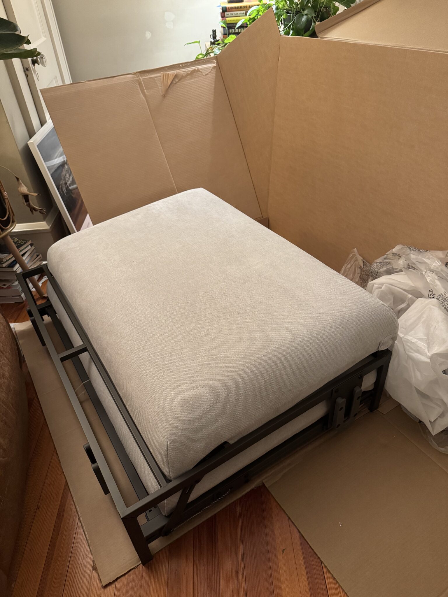 A light gray cushioned ottoman with a black metal frame sits partially assembled on a wooden floor, surrounded by cardboard packaging and plastic wrapping.