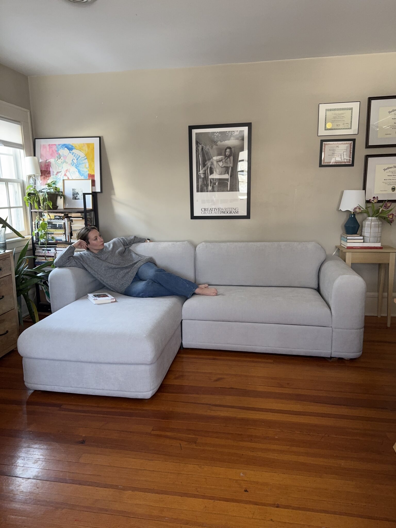 A person lounges on a light gray sectional sofa in a living room with wooden floors, wall art, framed certificates, books, and plants.