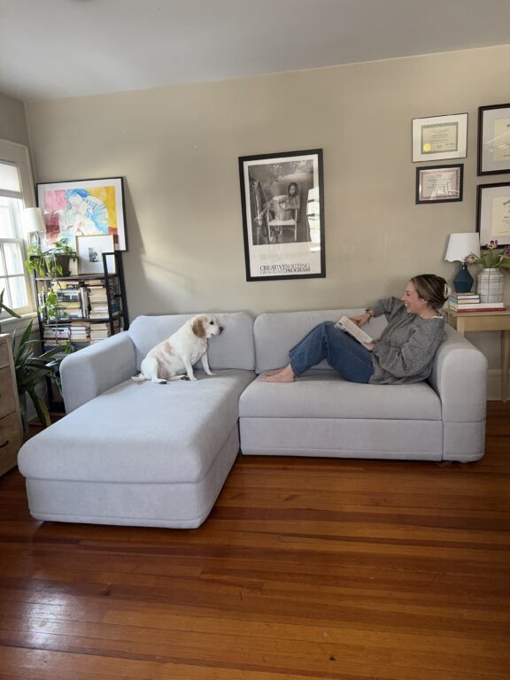 A woman sits on a light gray sectional sofa reading a book while a small white dog sits beside her; framed art and certificates hang on the wall behind them.