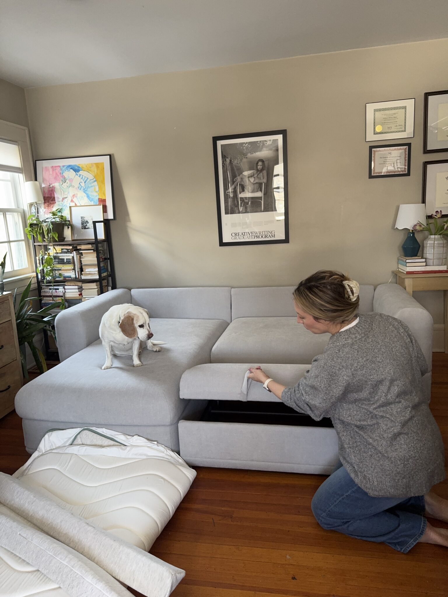 A woman kneels next to a light gray sofa, lifting the storage compartment under the seat, while a small white and brown dog sits on the sofa.