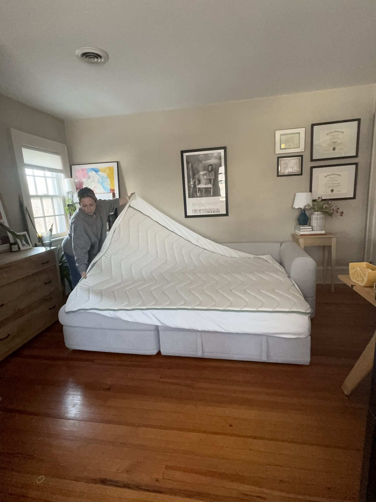 A person stands next to a pull-out sofa bed, lifting the mattress cover in a neatly arranged room with framed art and certificates on the wall.