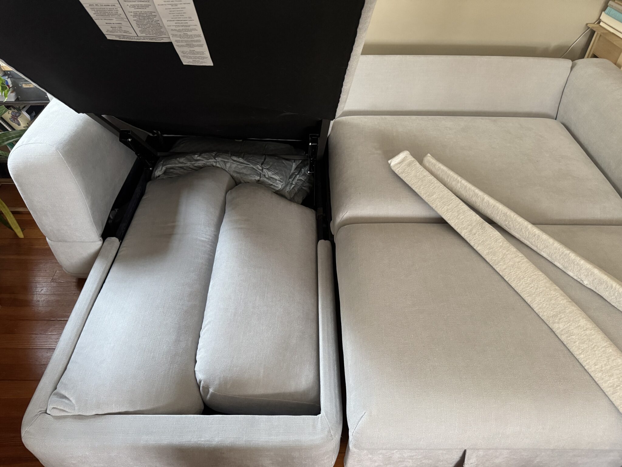 A light gray sectional sofa with an open storage compartment containing cushions; two long fabric-covered pieces sit on the adjacent seat.