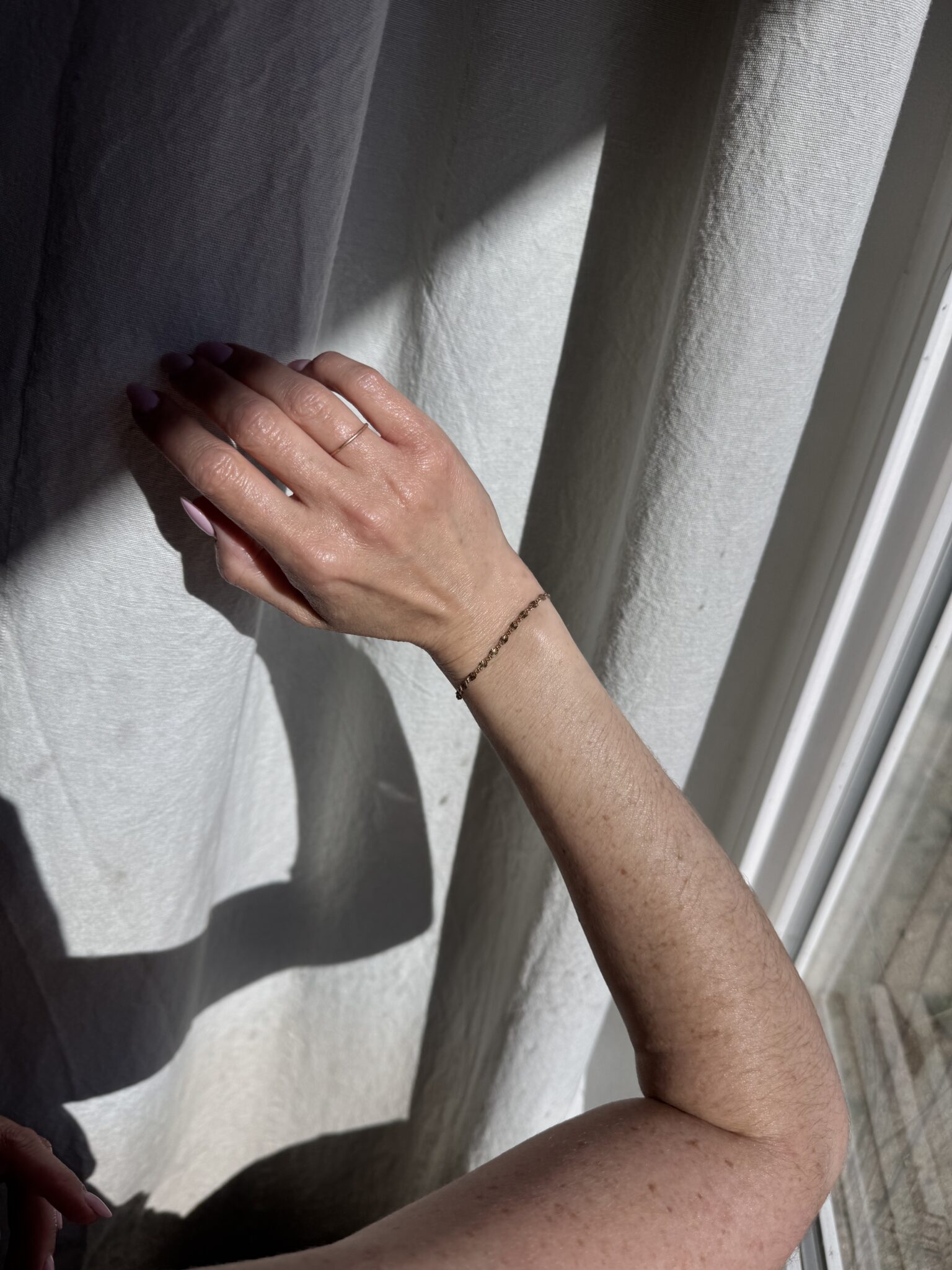 A person's left hand and forearm with a thin bracelet rest against a sunlit white curtain, casting a shadow.