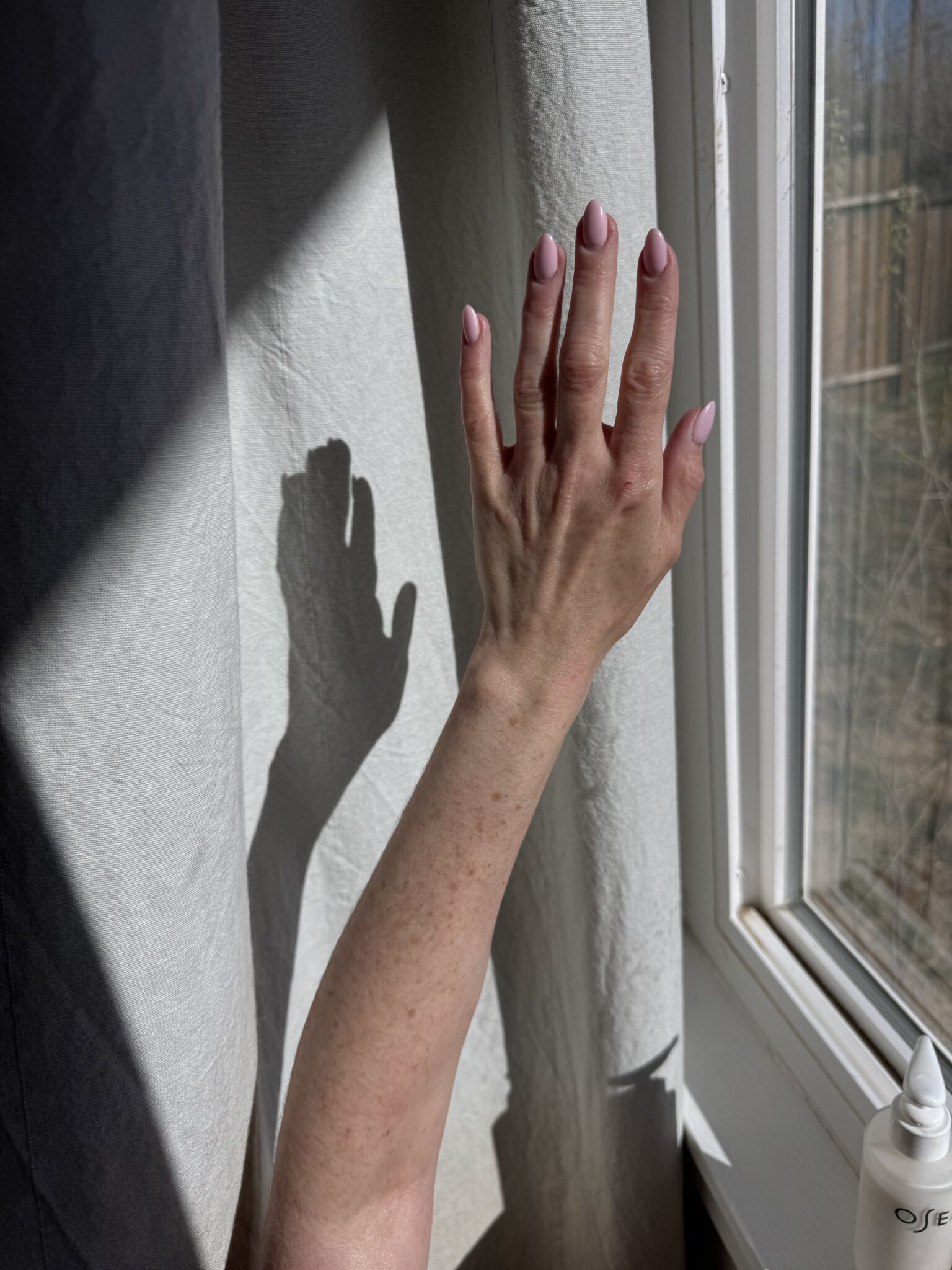 A hand with pale pink nails is raised toward a sunlit window, casting a shadow on a light-colored curtain. A bottle is partially visible in the bottom right corner.
