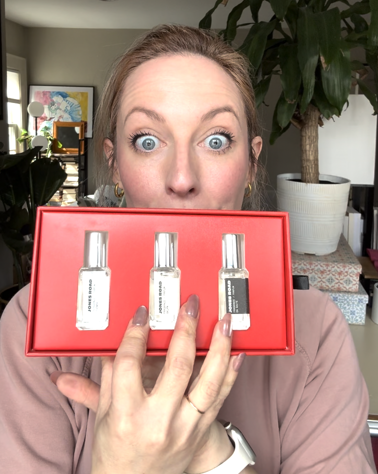 A woman holds up an Ilia red box containing three small bottles of perfume in front of her face, with a surprised expression.