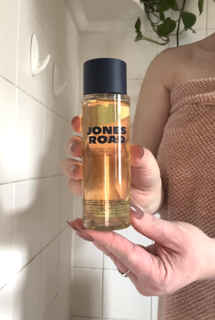 A person in a towel holds a clear bottle of Jones Road beauty product, reminiscent of Ilia’s minimalist style, in a tiled bathroom.