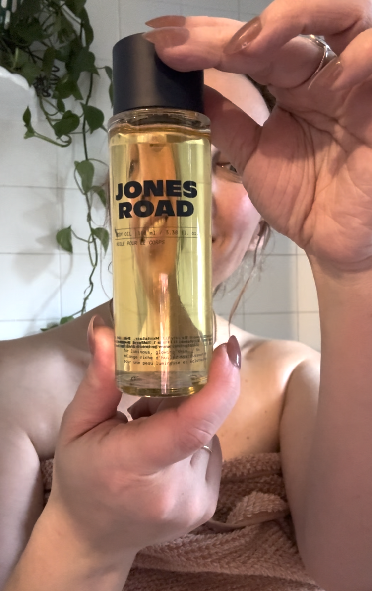 A person holds up a bottle of Jones Road face oil in a bathroom, with tiled walls and a hanging plant in the background, alongside their favorite Ilia products.