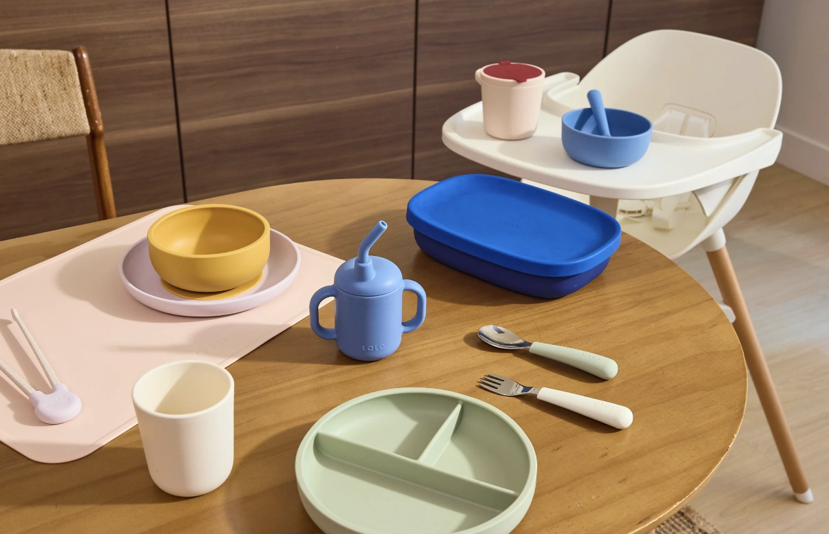 A wooden table set with colorful toddler dishware, utensils, a sippy cup, and a high chair in the background.