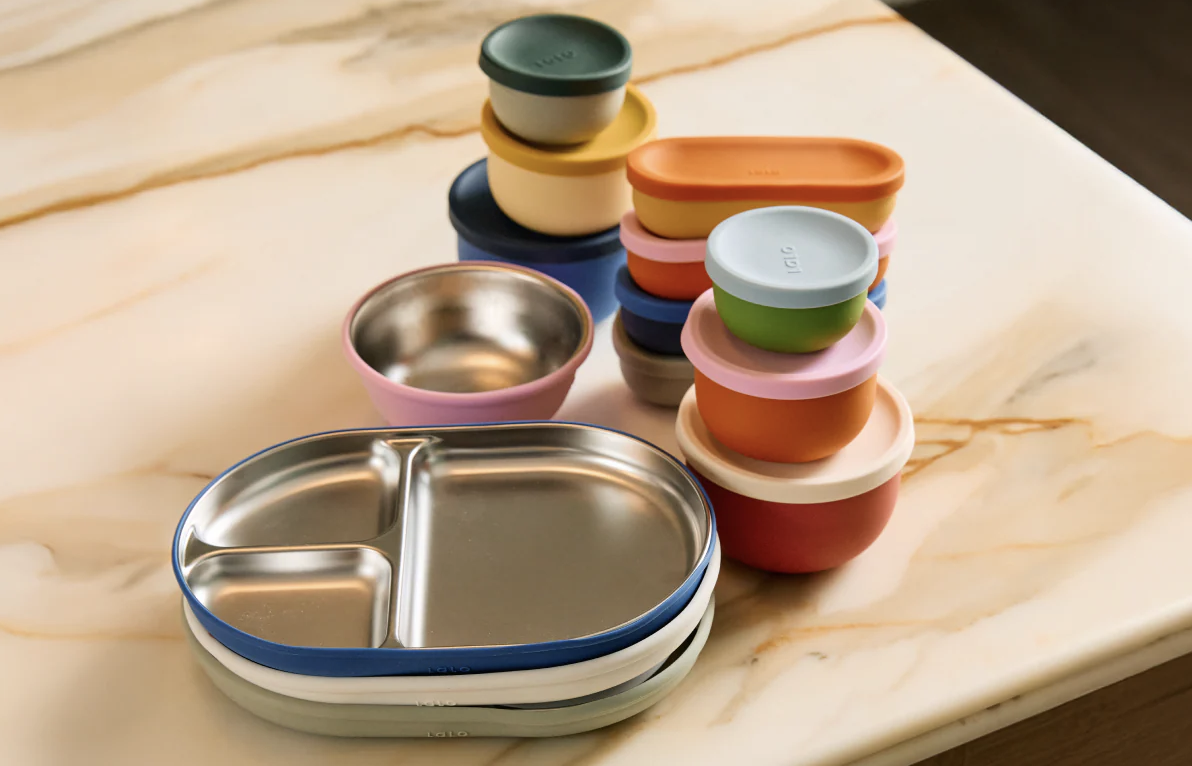 Assorted colorful silicone and stainless steel food containers and bowls are neatly stacked on a marble countertop.