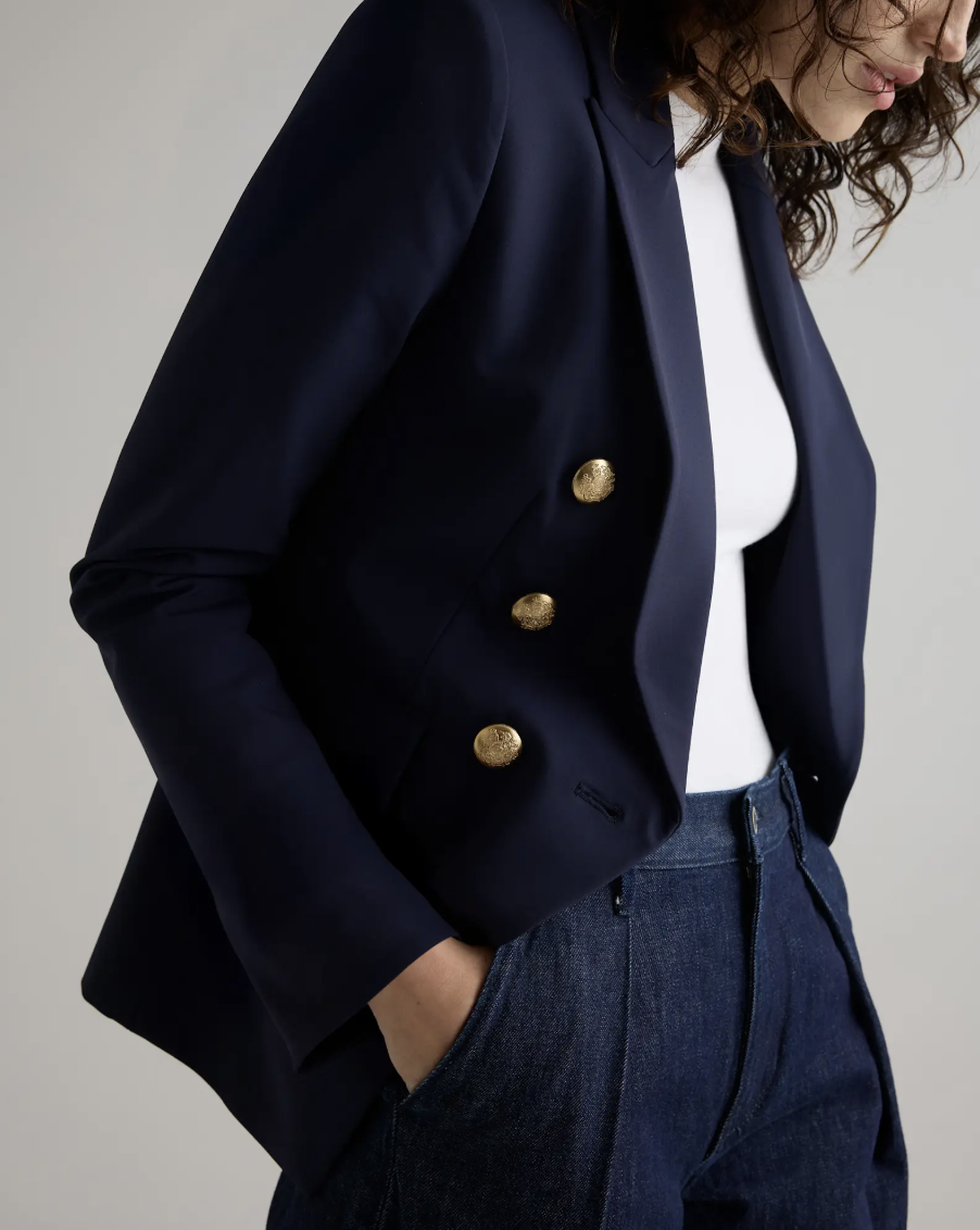 Person wearing a navy blazer with gold buttons, a white top, and dark blue jeans, with one hand in their pocket.